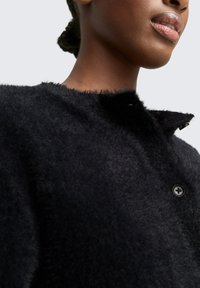 Schwarzer, flauschiger Mantel mit rundem Halsausschnitt, weicher Textur und Knopfschluss. Nahaufnahme, die das Detail des Kragens und den Stoff hervorhebt.