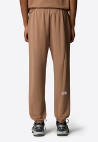 Lichtbruine trainingsbroek met een elastische tailleband, voorzien van een klein wit logo op het linkerpijpje en afgewerkte boorden. Gecombineerd met grijze sneakers.