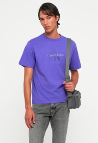 Mlad čovjek nosi ljubičastu Calvin Klein majicu, sive traperice i sivi crossbody torbu, stoji uz jednostavnu bijelu pozadinu.