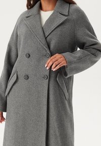 Manteau gris croisé en mélange de laine. Présente de grands revers, deux poches avant et des boutons noirs. Manches longues et amples.