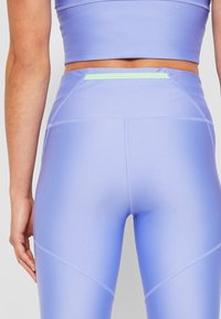 Detailní záběr osoby v levandulových sportovních legínách s vysokým pasem a odpovídajícím zkráceným topem, zaměřený na záda a boky ruky.