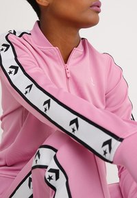 Pink athletic tracksuit featuring a zippered jacket with black and white patterned stripes along the sleeves. Smooth texture, fitted design.