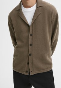 Homme portant un cardigan marron boutonné sur un t-shirt blanc à col rond et un pantalon noir, devant un fond uni.