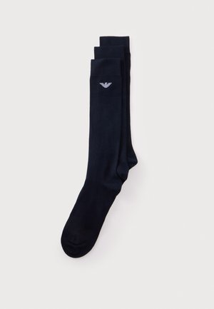 Une paire de chaussettes bleu marine foncé montant jusqu’aux genoux, ornées d’un petit logo blanc représentant un aigle, sur un fond clair uni.