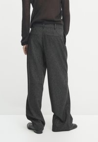 Person wearing loose-fitting dark gray pinstripe trousers and black shoes, standing against a plain light background, shown from the back.