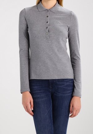 Frau trägt ein graues langärmliges Poloshirt mit Knöpfen und blaue Jeans, steht mit locker an den Seiten hängenden Armen.