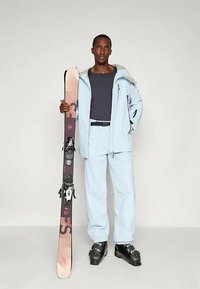 Mees helesinises suusajakas ja -pükstes, hoides ühte suuska, jalas mustad suusapüksid, seismas ühtlasel taustal.