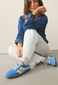 Blauwe suède sneakers met crèmekleurige accenten, een platte bruine zool en witte veters. Het ontwerp heeft een gladde afwerking en contrasterende kleurvlakken.