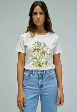 Mujer con camiseta blanca con estampado floral colorido y texto "SALSA JEANS", combinada con jeans de cintura alta en azul claro, posando frente a un fondo liso.