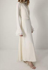 Femme portant une longue robe couleur crème avec des poignets évasés, des côtés plissés et un collier de perles superposé, debout devant un fond uni.