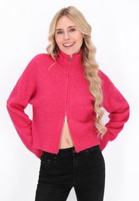 Maglione aderente e corto con zip di maglia rosa brillante, dotato di un alto colletto e maniche lunghe. Abbinato a jeans neri a vita alta.