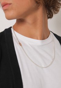 Collier en chaîne argentée au design plat et entrelacé, porté sur un t-shirt blanc et sous un cardigan noir. Texture lisse, détails minimalistes.