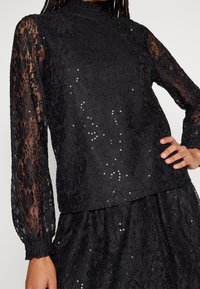 Haut en dentelle noir avec des manches longues, col montant, motif floral et paillettes. Comprend des poignets froncés et un design superposé.