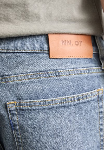 Niebieskie dżinsy mają skórzaną naszywkę z wytłoczonym napisem "NN. 07" na pasku i widoczne detale szwów na tylnej kieszeni.