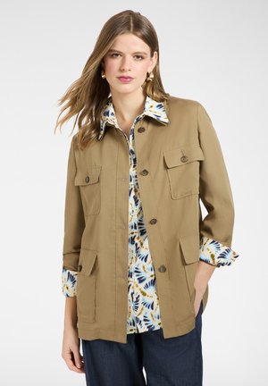 Frau trägt eine beige Jacke mit Knöpfen über einer gemusterten Bluse mit blauen und braunen Akzenten, kombiniert mit dunkelblauen Jeans, vor einem schlichten weißen Hintergrund.