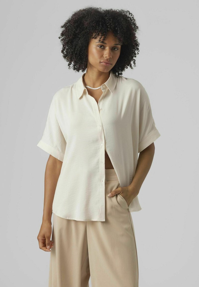 Camisa color crema con botones, fabricada en tejido ligero, con mangas cortas enrolladas y un corte relajado, combinada con pantalones de pierna ancha en color beige.