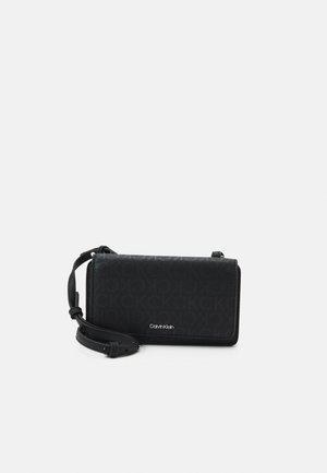 Černá crossbody taška Calvin Klein s monogramovým vzorem, nastavitelným popruhem a předním klopovým uzávěrem na bílém pozadí.