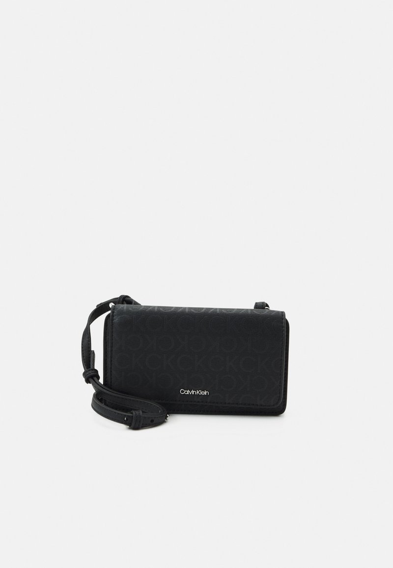 Czarna torebka crossbody Calvin Klein z monogramem, regulowanym paskiem i zapięciem na klapkę na białym tle.