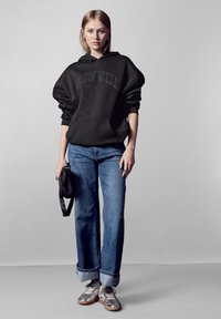 Felpa nera oversize con tasca frontale e logo, abbinata a jeans dritti azzurri chiari e sneakers argentate. Presenta polsini arrotolati.