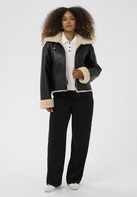 Čierna kožená bunda s krémovým ovčím golierom a manžetami, v kombinácii s bielou blúzkou s golierom a čiernymi širokými nohavicami. Jednoduché pozadie.