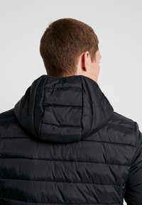 Doudoune noire avec capuche, présentant un design matelassé avec des coutures horizontales et une texture lisse et mate.