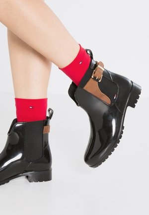 Jambes portant des bottines noires brillantes avec des détails de boucle marron et des chaussettes rouges vives avec une petite broderie de logo.