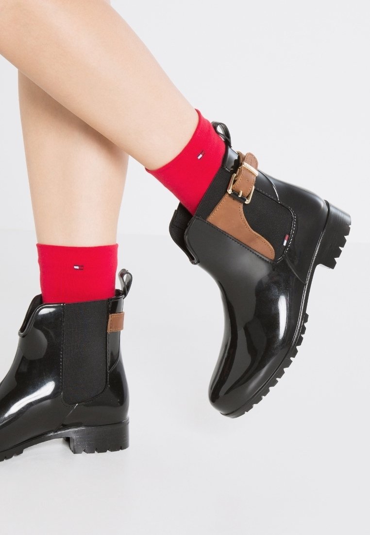 Jambes portant des bottines noires brillantes avec des détails de boucle marron et des chaussettes rouges vives avec une petite broderie de logo.