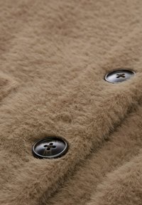 Beige textiel met een zachte, fuzzy uitstraling, met twee zwarte knopen met een ontwerp van vier gaatjes.