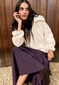 Zgostljeni pleteni pulover z razširjenimi rokavi, skupaj z bordo karirasto krilo. Obleko zaokrožijo rjavi gležnjarji. Leseno in kamnito ozadje.