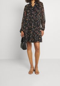 Zwarte bloemenjurk met lange transparante mouwen, geplooide rok en een V-hals. Gecombineerd met bruine hoge sandalen en een zwarte gevlochten handtas.