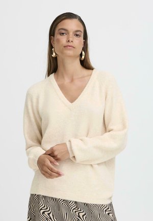 Helles beige V-Ausschnitt-Pullover mit langen Ärmeln und weicher Textur, kombiniert mit einem gemusterten Rock, der gewellte schwarze Linien auf einem beigen Hintergrund zeigt.