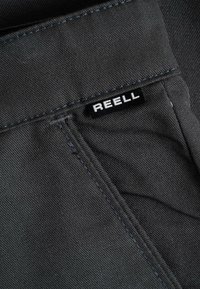 Black fabric close-up showing a stitched pocket corner with a small black and white REELL brand label attached.