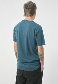 Türkises Baumwoll-T-Shirt mit kurzen Ärmeln, rundem Hals und flachen Nähten. Es hat eine glatte Textur und ist in eine dunkle Hose gesteckt.