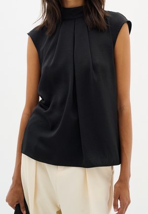 Femme portant un chemisier noir sans manches avec des plis plats sur le devant et un col montant, associé à un pantalon large couleur crème, tenant une pochette noire.