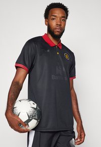 Zwarte poloshirt met rode kraag, voorzien van een logo en voorbranding. Model houdt een zwart-wit gestreepte soccerbal vast.