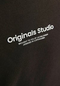 Tessuto nero con superficie testurizzata che presenta testo stampato bianco: "Originals Studio" e "PORTATO A TE DA JACK&JONES® PROGETTATO A COPENAGHEN."