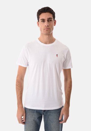 Giovane uomo che indossa una semplice T-shirt bianca a maniche corte con un piccolo logo rosso e nero sul petto a sinistra, abbinata a jeans blu.