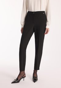 Czarne dopasowane spodnie z elastycznym pasem, z przednim guzikiem, plisami i delikatnym połyskiem. Zestawione z lśniącymi czarnymi klapkami na obcasie.