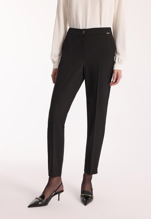 Czarne dopasowane spodnie z elastycznym pasem, z przednim guzikiem, plisami i delikatnym połyskiem. Zestawione z lśniącymi czarnymi klapkami na obcasie.