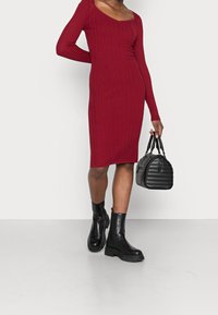 Robe en tricot côtelé bordeaux avec manches longues et encolure carrée, associée à des bottines en cuir noir et un sac matelassé.