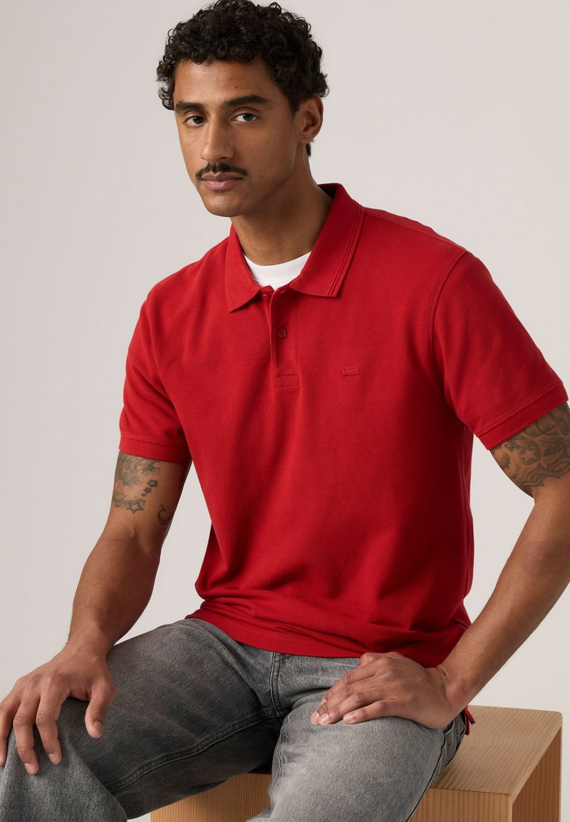 Homme aux cheveux bouclés et tatouages assis sur un bloc de bois, portant un polo rouge et un jean gris sur un fond uni.