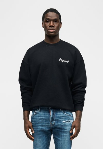 Νεαρός άνδρας που φοράει μαύρο φούτερ Dsquared2 και μπλε φθαρμένο τζιν, στέκεται μπροστά σε απλό φωτεινό φόντο.