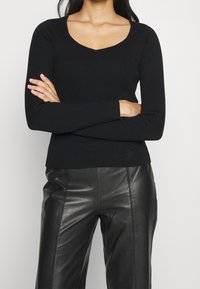 Svart ribbad långärmad topp med V-ringning och figurnära silhuett, kombinerad med svarta läderbyxor som visar en jämn textur och kontur.