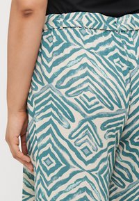 Lightweight fabric pants with a teal and white zigzag pattern, featuring a belted waist and side pockets. Smooth texture, relaxed fit.