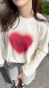Sweatshirt à manches longues couleur crème avec un grand motif de cœur rouge dégradé au centre, associé à un short en denim et un sac à main noir.