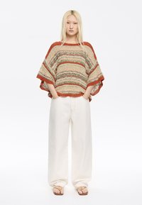 Strickponcho in mehrfarbigen Streifen mit lockerer Passform, rundem Ausschnitt und dekorativen Bordüren. Kombiniert mit weiten, weißen Hosen.