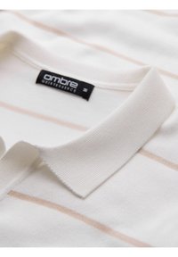 Bílá polo košile s texturovaným límcem, měkkou tkaninou a tenkými horizontálními pruhy v jemně broskvové barvě. Obsahuje černou značkovou etiketu.