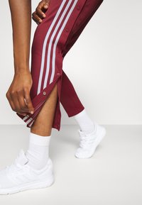 Bordeauxfarbene Sporthosen mit Druckknopfverschlüssen an den Seiten und drei weißen Streifen. Getragen mit weißen Turnschuhen und Socken, die eine strukturierte Passform bieten.