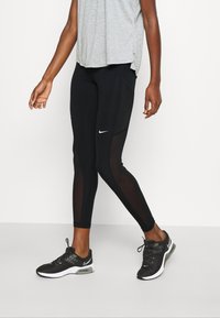 Svarta leggings med nätpaneler, Nike-logotypen fram. Kombinerat med en ljusgrå topp och svarta träningsskor. Slät textur.