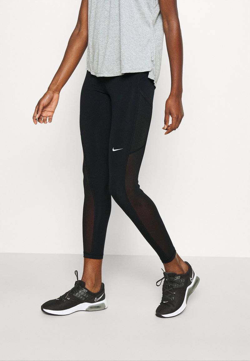 Svarta leggings med nätpaneler, Nike-logotypen fram. Kombinerat med en ljusgrå topp och svarta träningsskor. Slät textur.